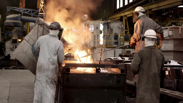 A team of foundry workers uses a transfer ladle to pour the flaming, smoking, glowing molten steel into a mold at an industrial workshop.