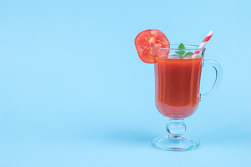 Glass glass with tomato juice, with a cocktail tube and mint leaves on a blue background.