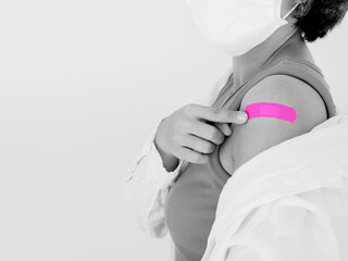 Vaccinations concept. Vaccinated woman wearing medical face mask pointing on pink bandage plaster on her shoulder after vaccination treatment isolated on white with copy space, black and white style.