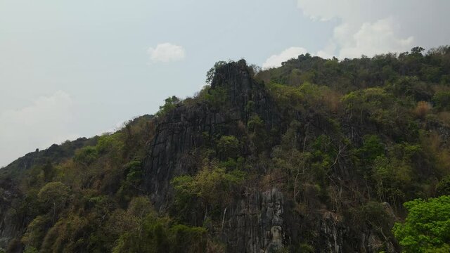 4K Vertical descending aerial footage of a limestone steep cliff that extruded out of a mountain with green vegetation growing all over surface seen during the summer in Thailand.