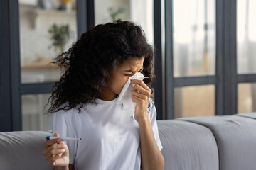 Cold and flu concept. Sick exhausted African American woman with runny nose, headache and fever is blowing out snot using napkin. Home treatment