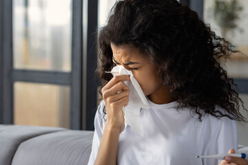 Fototapeta na wymiar Home treatment. Sick exhausted African American woman with runny nose, headache and fever is blowing out snot using napkin. Cold and flu concept