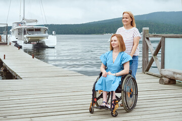 A woman in a wheelchair walks with her sister or her friend on a wooden pier by the sea