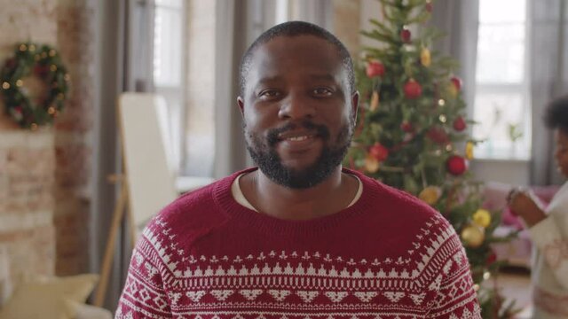 Portrait of cheerful Afro-American man smiling and looking at camera while his wife decorating Christmas tree at home in background