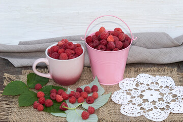 There is a cup and a bucket of raspberries on a jute fabric.