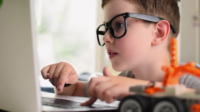 Side view of smart boy in eyeglasses programs a robot car at a laptop at home. Kid learning coding and programming. Robotics, Science, Mathematics, Engineering, Technology, Education. Moving image.
