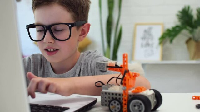 Clever boy in eyeglasses programs a robot car using a laptop at home. The child is learning coding and programming. Robotics, Science, Mathematics, Engineering, Technology. STEM education.
