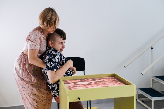 Children with disability have mobility problems. Mother carrying boy with cerebral palsy Woman assists and transport on chair. Rehabilitation center, mother with teenage person at home