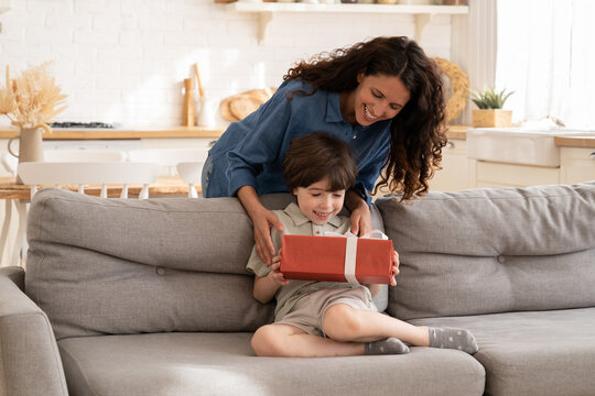 Excited little boy receiving birthday present from mum sitting on sofa in living room. Children holiday celebration concept with loving mother giving gift to adorable preschool son congratulating kid
