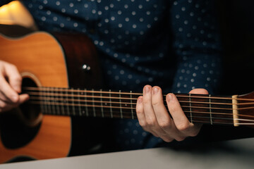 Close-up front view of unrecognizable guitarist male playing acoustic guitar sitting at desk in dark living room, selective focus. Creative musician enjoying leisure activity in apartment.