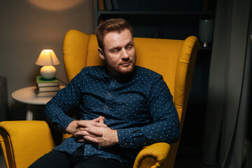 Medium shot portrait of confident handsome man in stylish shirt sitting comfortably in luxurious yellow armchair with arms crossed, in dark cozy living room with modern interior, looking at camera.