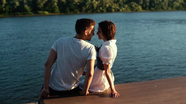 Back view of young man trying to kiss him lover sitting by river in sunny day, girlfriend refuse. First date in city park, lady rejects kiss from her suitor, rear view. Concept of unrequited love.