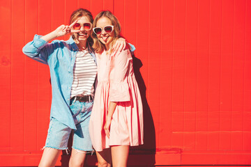 Two young beautiful smiling hipster female in trendy summer  clothes.Sexy carefree women posing on street background near red colorful wall.Positive models having fun and hugging. Cheerful and happy