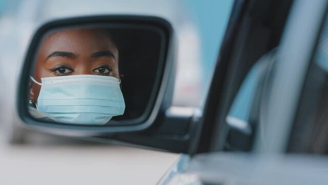 Reflection in car mirror beautiful young african american woman driver black mixed race girl with stylish makeup wears medical protective mask on female face sitting looking in automobile auto salon
