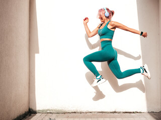 Fitness smiling woman in green sports clothing with pink hair. Young beautiful model with perfect body.Female jumping and jogging in street near white wall.Listening music in wireless headphones