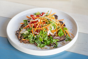 Fresh summer salad made of roasted beef meat slices, lettuce, grated carrot, sweet pepper and onion decorated with sesame seeds served on white round plate on striped wooden background at restaurant
