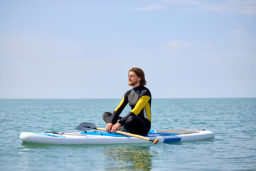 Naklejka premium Athlete surfer mpractice yoga exercise on sup board at sea in relaxing day, meditating alone while sitting on surfing board, keep calm. yoga is meditation and healthy sport concept