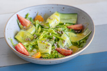 Refreshing summer salad made of raw green cucumber slices, lettuce and red strawberries decorated with sunflower seeds and sauce served on plate on striped wooden background at restaurant. Horizontal