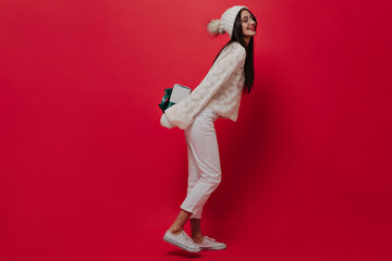 Fototapeta na wymiar Full-length photo of trendy young brunette in white jeans, sneakers, knitted jersey and hat, smiling with closed eyes, preparing gift box isolated on red plain background 