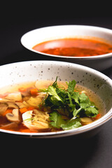 Chicken soup with vegetables amd mushrooms, fresh parsley and dill on top. Served in a white bowl over black background