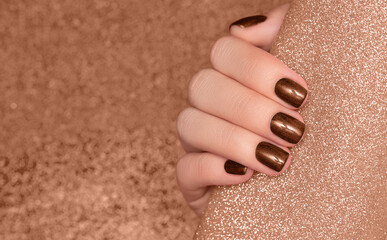 Female hand with brown nail design. Glitter brown nail polish manicure. Woman hand on glitter beige fabric background.