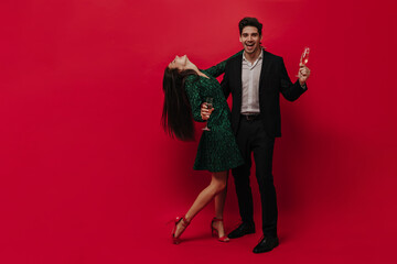 Engaging young dark-haired man in black suit and white shirt smiling, celebrating with glass of champagne and holding her girlfriend against red background. Lady wearing green dress and bright shoes