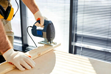 close-up construction worker cuts wooden board with an electric jigsaw while renovating house, cropped male in gloves repairing using electric tools, equipment