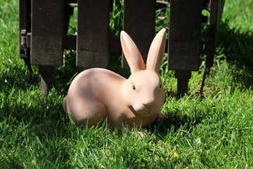 Creepy ceramic light brown garden decoration rabbit looking directly in camera next to plastic fence surrounded with uncut grass on warm sunny spring day