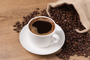 Coffee cup and saucer on wooden background with roasted coffee beans in a burlap sack.