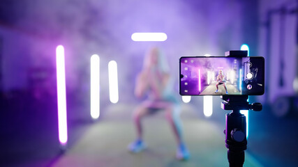 Professional cute female fitness trainer video record exercise on smartphone surrounded by neon lights