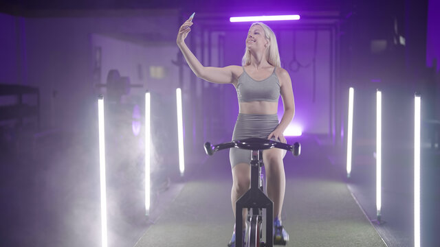 Instagramer Fitness Girl Making A Selfie Photo On Gym Bicycle In Neon Lights