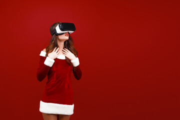 Obraz premium Amazed young Santa girl in Christmas dress looking in headset pointing hands aside isolated on red wall background. 
