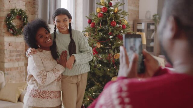 Selective focus shot of happy Afro-American mother and daughter embracing and smiling by Christmas tree at home as father taking picture of them with smartphone