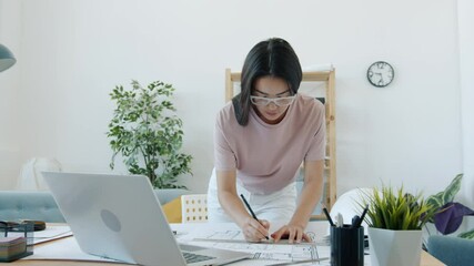 Young female architect is working at building plan drawing on blueprint and using laptop at home. Professionals and modern technology concept.