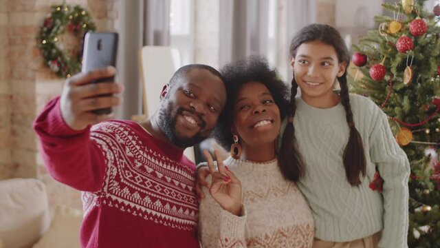 Cheerful Afro-American family smiling and posing together for smartphone camera while taking selfie by Christmas tree at home