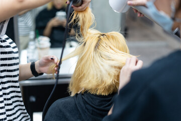 Two Asian professional female hairdressers blowing a young Asian female customer's hair with a hair dryer while sitting on chair and using a cell phone in front of a blurred mirror in a hair salon.