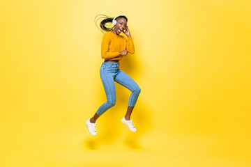 Fototapeta na wymiar Happy energetic young African American woman wearing headphones listening to music and jumping in yellow isolated studio background