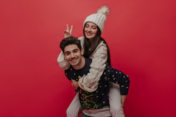 Couple of young joyful brunette-haired people, having fun, smiling and posing in cute winter outfits isolated on red background 