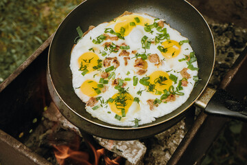 Frying pan of fried eggs, bacon and organic greens, green grass on the background, cooked on the grill on a summer sunny day, summer breakfast, eating in nature - the concept of a healthy lifestyle