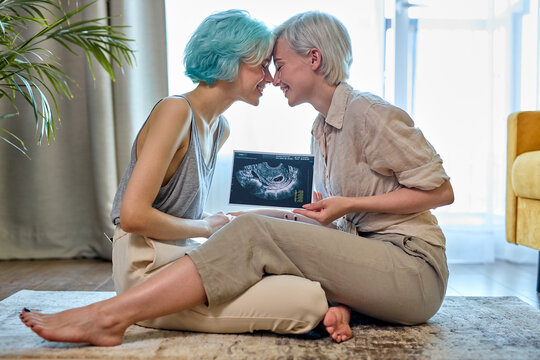 Lgbt couple sit on living room floor together kissing and hugging after news about pregnancy, waiting to be mothers, one of them is pregnant, baby expectant. at home side view portrait, copy space