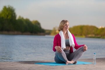 Mature Caucasian Blond Woman During Yoga Practice With Eyes Closed on Blue Mat At Water Shore Outdoor.