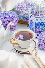 A visual for content. Still life in vintage style. A mug with a drink, an old book, a cage, candles and hydrangea flowers in the garden on a white wooden table. The concept of a tea ceremony.