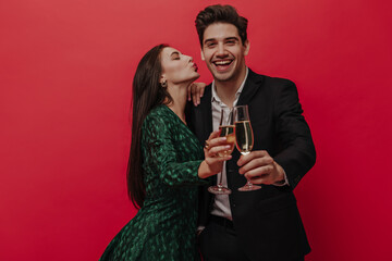 Fototapeta na wymiar Cute young brunette-haired lady in green dress holding glass of wine and trying to kiss her husband wearing white shirt and black suit. Pair posing isolated on red background 