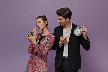 Engaging young man in white shirt and black suit showing time to his girlfriend. Lady wearing pink dress, making makeup against violet background 