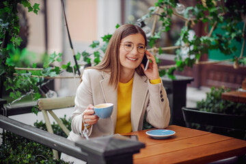 Smiling young woman in yellow sweater sitting on terrace of cafeteria, holding cup of coffee and talking on a phone with friend.