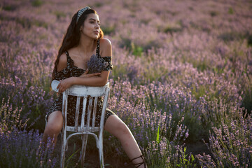 Beautiful girl in a black dress sits on a wooden chair and poses in the middle of a lavender field.