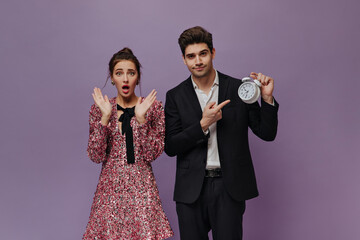 Surprised young lady with brunette hair posing in pink mini dress near pretty boy wearing black suit and showing time on clock. Pair standing against violet background 