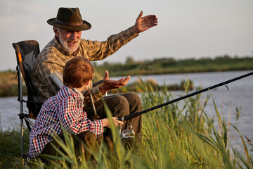 Grandfather and kid boy together fishing in the evening at sunset time in summer day on river in countryside, side view on caucasian multi-generation family, elderly man teach child boy to fish