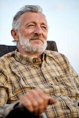 Handsome Senior Man In Checkered Shirt Sit On Chair In Nature, Having Rest Alone, Thinking, Smiling, In Contemplation Of Countryside Landscapes, Portrait Of Gray Bearded Male Relaxed