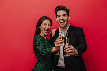 Cheerful young pair of people in holiday outfits, smiling, holding glasses with champagne and looking into camera isolated on red background 